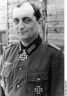 Black-and-white portrait of a man wearing a military uniform with an Iron Cross displayed at his neck, his dark hair is combed back.
