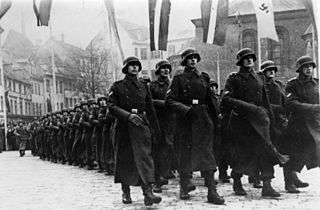 a black and white photograph of soldiers in German uniform and greatcoats marching in a column