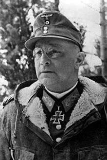 A black-and-white photograph of a man with glasses wearing a military uniform underneath a fur collared coat, field cap and neck order in shape of an Iron Cross.