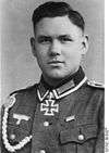 The head of a young man, shown in semi-profile. He wears a military uniform with a military decoration in shape of an iron cross displayed at the front of his shirt collar. His hair is parted and combed to back.