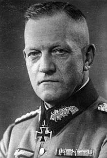 Black-and-white portrait of a determined looking man wearing a military uniform with an Iron Cross displayed at his neck, his hair is combed back.