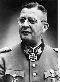 A man wearing a military uniform, glasses and neck order, in the shape of a cross. He has short hair that is combed back and a determined facial expression.