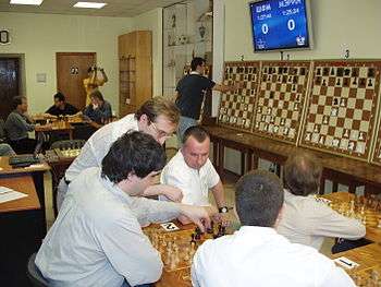 Photo of a room with several chessboards on tables, groups of men playing around two of them. On the wall, several large demonstration chessboards