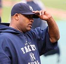 A dark-skinned man in a navy-blue sweatshirt and baseball cap grabs the brim of his cap with his left hand.