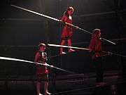 Two tightrope walkers with balance poles support a third who stands on a bar supported by the initial two walkers' shoulders.