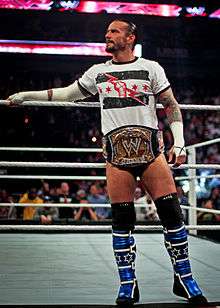A man stands on a wrestling ring apron, a golden belt with the WWE insignia around his waist.