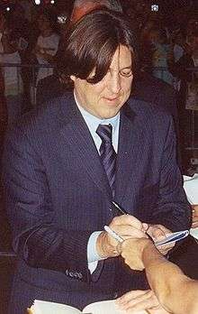 A man wearing a suit looks down at the peace of paper he is signing for the person who is out of shot.