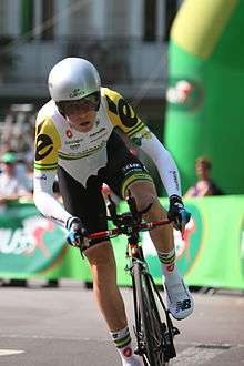 A road racing cyclist wearing a predominantly white and black skinsuit with green and gold trim.