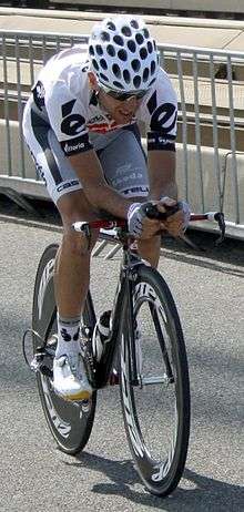 A road racing cyclist in a mostly white jersey with black trim riding a bicycle with a solid disc rear wheel.