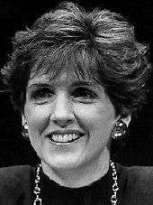 A black and white image of a pale-skinned woman in her early forties, with fairly dark hair with a swept hairdo covering part of her ears, smiling, wearing solid metal earrings and a metal chain necklace and a dark top.