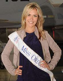 A blonde woman in a purple dress and tan jacket wearing a Miss Oregon pageant sash