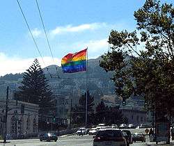 A color photograph of a large Gay Pride flag flying at the intersection of Market and Castro Streets and the hills of San Francisco in the distance