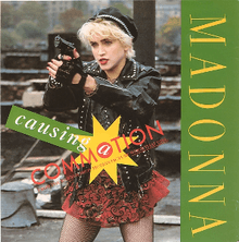 Madonna with short cropped blond hair is pointing towards somebody while holding a gun. She is wearing a red skirt and black jacket and gloves.