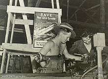 Black-and-white photograph of two soldiers working with a lathe. A poster behind them depicts a ship with hospital markings sinking by the bow and is captioned with "SAVE for the brave" and "Let us avenge the Nurses".