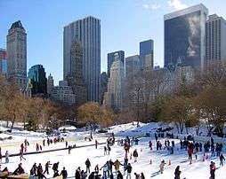 An outdoor skating rink with many people on the rink. There are skyscrapers in the background. This is the Wollman Rink in Central Park.
