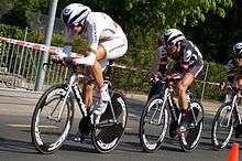 Three cyclists in aerodynamic skinsuits riding single-file. They are crouched low over their bicycles. The first in the line wears a special jersey, predominantly white with yellow, green, and red stripes in the middle. The others wear identical black and white jerseys with red trim.