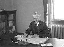 A balding white man in a three piece suit sits at a desk, pen in hand, with documents laid out before him.
