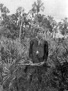 A black and white photograph of a Seminole man wearing traditional Seminole smock and vest, holding a rifle standing among palmettos, and staring at the viewer