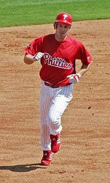 A man in white pants, a red baseball jersey with "Phillies" on the chest, and a red batting helmet with "P" on it runs on a dirt surface.