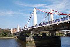 Suspension bridge crossing a wide river, under blue skies with clouds. The main cables are red and the towers white.
