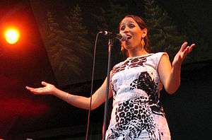 A woman wearing a black-and-white printed dress, standing behind a microphone with her arms extended.
