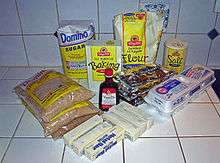 An arrangement consisting bags of brown sugar, white sugar, flour and chocolate chips; boxes of baking soda and salt; a carton of eggs, wrapped sticks of butter and a bottle of vanilla extract on a kitchen counter