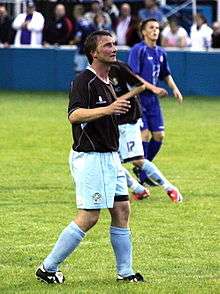 A man with dark hair who is wearing a black top, light blue shorts and light blue socks. He is standing on a grass field.