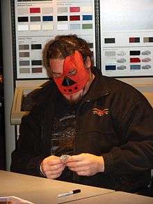 Large adult male wearing a black shirt and jacket, as well as a red mask over his face with shirt brown hair.