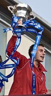 A man with dark hair is wearing a red jacket. He is holding aloft a trophy.