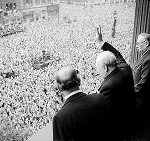 A balding man with a cigar in his mouth waves from a balcony to large crowds below him that fill the square.
