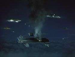 The scale model of an object that resembles an aircraft carrier hovering at high altitude emits fire and smoke. It is under missile attack from rotating, circular alien spacecraft surrounding it. The setting is night.