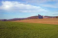 A landscape with a field and front and mountains showing fall color in the distance. At the right is a red barn with three blue silos in front and two older ones in back.