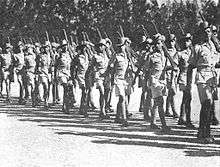 Soldiers in colonial-era military uniform march past, rifles shouldered.