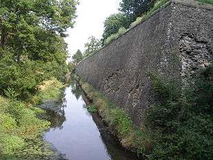 Photo shows an old stone fortress wall about 5 meters tall at right, a wet ditch in the center and trees at left.