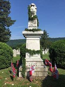 A stone Confederate monument decorated in an evergreen garland and surrounded by Confederate flags implanted in the ground beneath it.