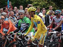 Several cyclists sit on their bicycles. In the center of the image are two, wearing solid yellow and green jerseys. Spectators watch in the background.