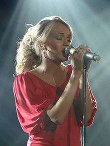 A young woman with blonde hair in a red top, holding a microphone and singing with her eyes closed.