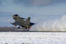 Three-quarter hind view of grey jet aircraft taking off in snowy environment.  Its engine is kicking up snow on the ground