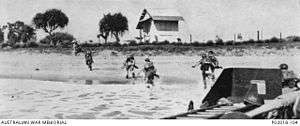 A black and white photograph showing soldiers moving from a landing craft on to a beach. In the foreground there is a tall wooden house on a ridge line