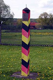 A red/black/yellow striped square-shaped pole with a metal spike on the top side. A metal plaque with an emblem and the words "Deutsche Demokratische Republik" is visible on one face.