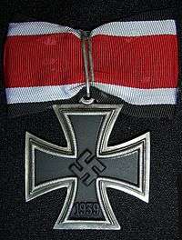 A silver framed black cross that has arms which are narrow at the center, and broader at the perimeter. In the middle of the cross is a swastika, an equilateral cross with its arms bent at right angles. On the lower arm of the cross are the number 1939 engraved. The cross is connected to a ribbon with a silver clip. The ribbon has a red central stripe, flanked in white and with a black edge stripe.