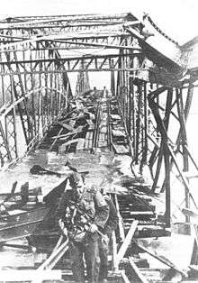 a black and white photograph looking along a damaged steel girder bridge from one end