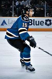Hockey player in blue and black uniform. He extends his stick to his right, and faces to the right of the camera.