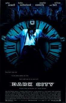 A black poster. Above reads the lines: "Rufus Sewell", "Kiefer Sutherland", "Jennifer Connelly", "and William Hurt". In the center, against a black background, a man wearing a blue jacket is rested against an upright clock with Roman numerals as big as him; the setup cast in a blue tint. His arms are outspread, and his head is tilted back with his mouth agape. Behind the man and the clock is a dark city skyline. Below them is the tagline, "They built the city to see what makes us tick. Last night one of us went off." Below the tagline is the film title, "Dark City".