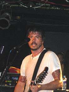 A bearded man wearing a white shirt plays guitar in front of a microphone.