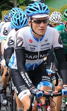 A road racing cyclist wearing a white jersey with black shorts, accented with blue argyle trim and a distinctive accented e on the shoulder. Several cyclists are behind him, including two teammates, ride behind him.