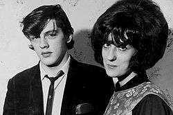 A young man and woman, in 1960s fashion, stand for a monochrome photograph. The man has a neutral expression on his face, the woman has a slight smile.