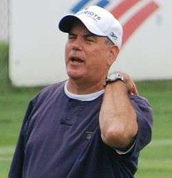 Candid photograph of Pees wearing a blue t-shirt and white baseball cap bearing the New England Patriots logo on a football field