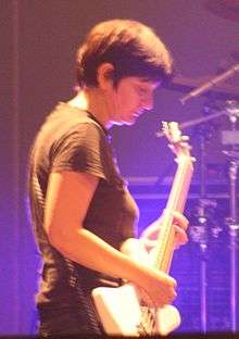 A woman performing live onstage with a bass guitar against a blue backlight.
