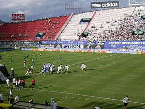 Two football teams, one in green and the other in white, preparing to start a match in a stadium with red and white seating sections, with more people in the latter; the sky is cloudy and blue.